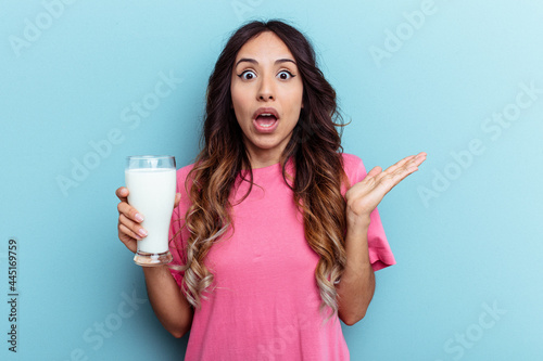 Young mixed race woman holding a glass of milk isolated on blue background surprised and shocked.
