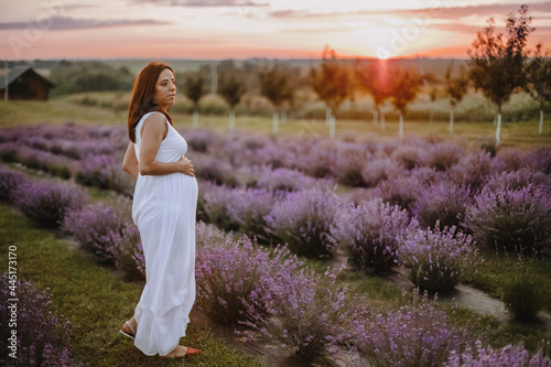  View in full height of happy woman expecting newborn child, standing at field with violet lavender and dreaming on sunset. Future mother looking to side, dressed in white outfit. Concept of pregnancy