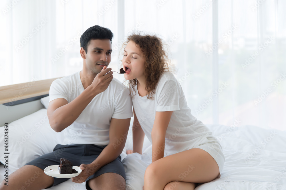 A man is sitting on the bed feeding the woman a chocolate cake. A man is sitting on the bed feeding the woman a chocolate cake.