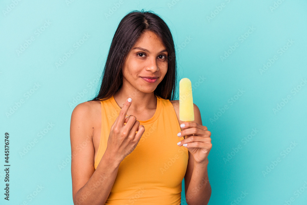 Young latin woman holding ice cream isolated on blue background pointing with finger at you as if inviting come closer. Young latin woman holding ice cream isolated on blue background pointing with finger at you as if inviting come closer.