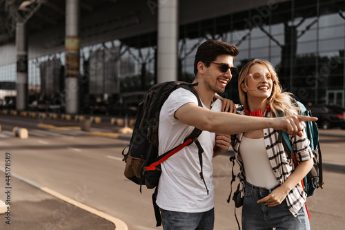 Travelers poses near airport in good mood. Man in white tee and woman in plaid shirt smiles and talking, holding backpacks.