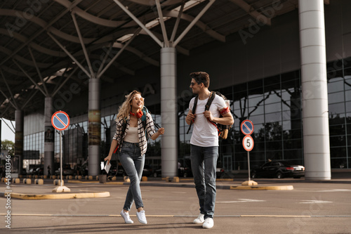 young woman in sunglasses and brunette man in jeans walking near airport. Happy girl in headphones and her boyfriend travels and poses with backpacks.