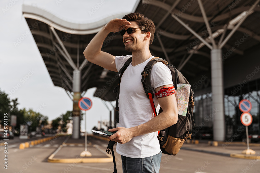 Fototapeta premium Stylish man in white tee and jeans smiles widely and looks into distance. Traveler with backpack holds passport near airport.