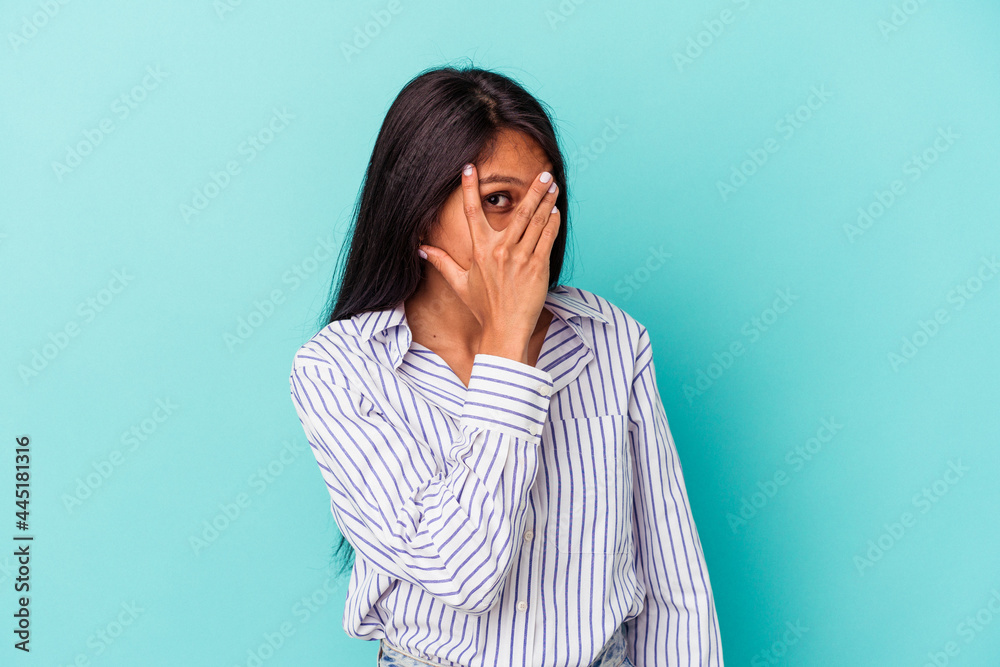 Young latin woman isolated on blue background blink at the camera through fingers, embarrassed covering face.