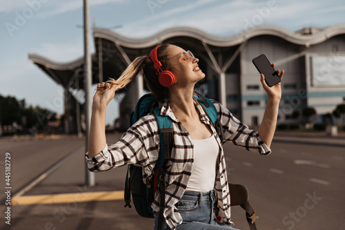 Charming blonde girl in sunglasses, red headphones listens to music. Pretty female tourist holds phone and backpack. Attractive woman in plaid shirt poses near airport.