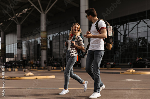 Joyful woman and man in white tees and jeans walks and talks near airport. Full-length portrait of travelers with backpacks.