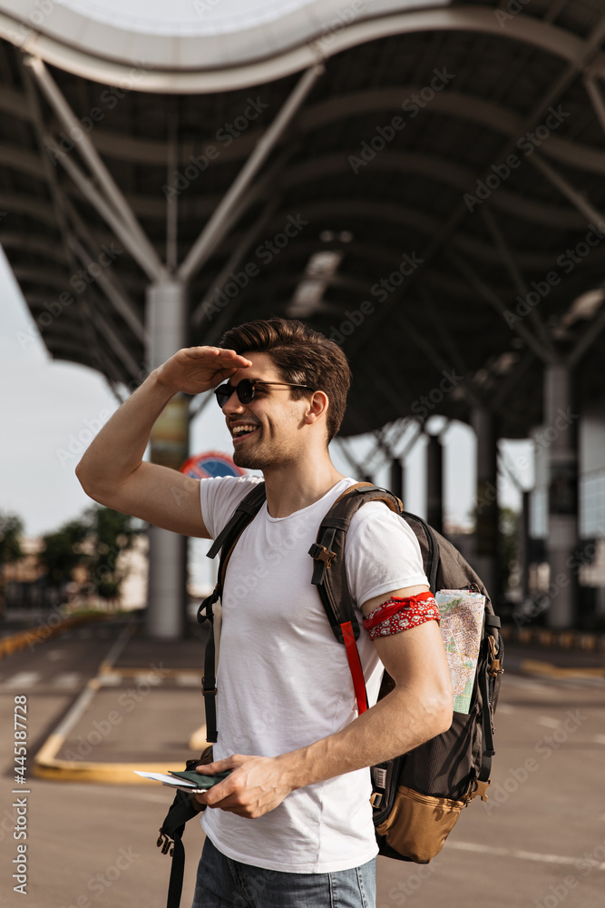 Portrait of cool man in white tee looking into distance and smiling. Brunette guy in sunglasses smiles and holds passport near airport. Portrait of cool man in white tee looking into distance and smiling. Brunette guy in sunglasses smiles and holds passport near airport.