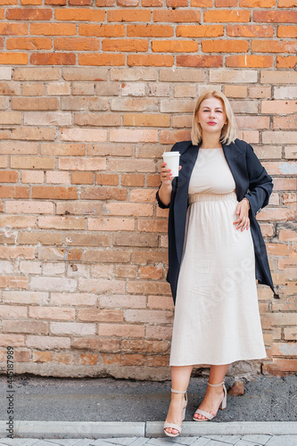 Happy beautiful young woman in a dress and jacket stands by a brick wall and drinks coffee
