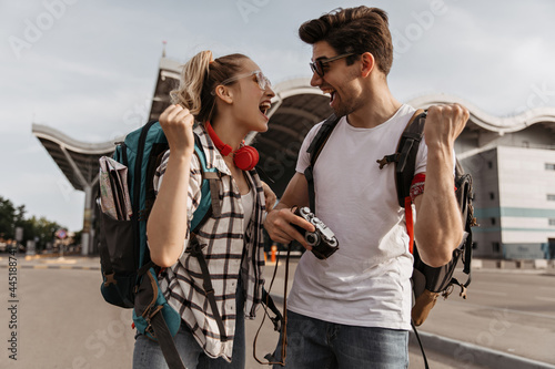 Young man and woman rejoice near airport. Blonde girl and brunette guy pose with backpacks outside. Make traveler holds retro camera.