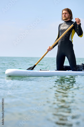 Male in wetsuit standing on paddle board at sea on blue water. Young male model on summer holidays vacation travel. Portrait of bearded guy holding paddles in hands, looking focused and confident