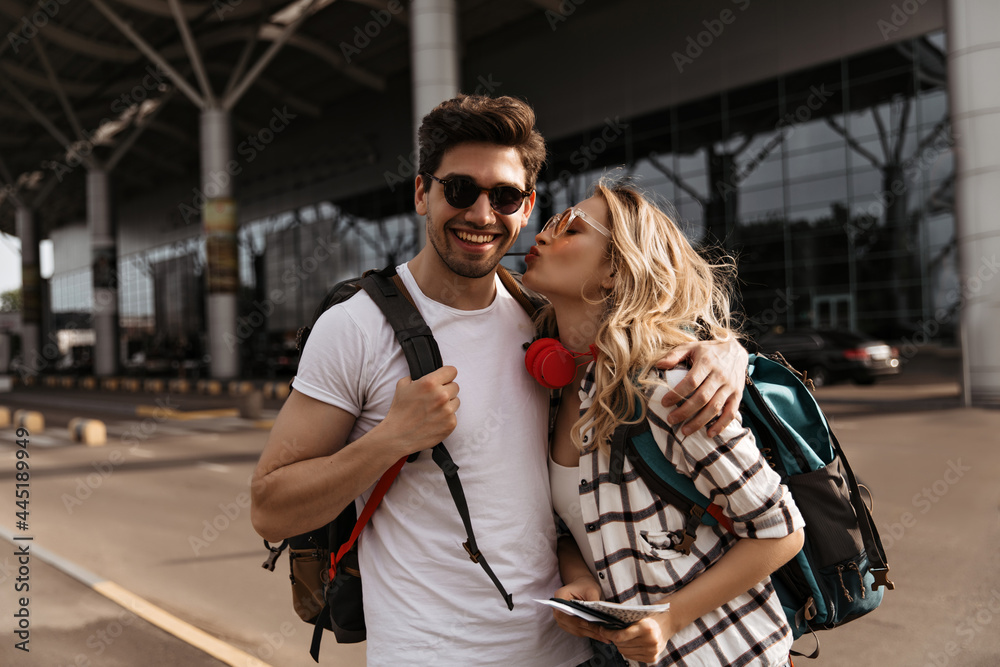 Naklejka premium Curly woman kisses her boyfriend near airport. Attractive girl in plaid shirt and brunette man in white tee poses in good mood and holds backpacks.