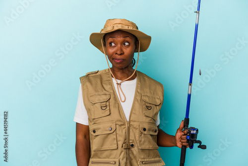 Young african american fisherwoman holding rod isolated on blue background confused, feels doubtful and unsure.