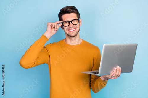 Photo of pretty attractive young man wear yellow sweater arm spectacles smiling holding modern gadget isolated blue color background