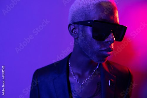 Portrait photo of a dark-skinned guy with a beard. Dressed in a black jacket and sunglasses. He has metal jewelry on his neck. Purple and pink light photo