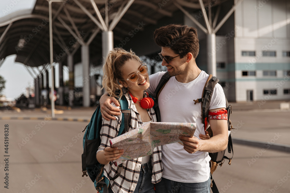 Fototapeta premium Cheerful woman and happy man in white tee-shirts smile and talk near airport. Travelers with backpacks hold map near airport.