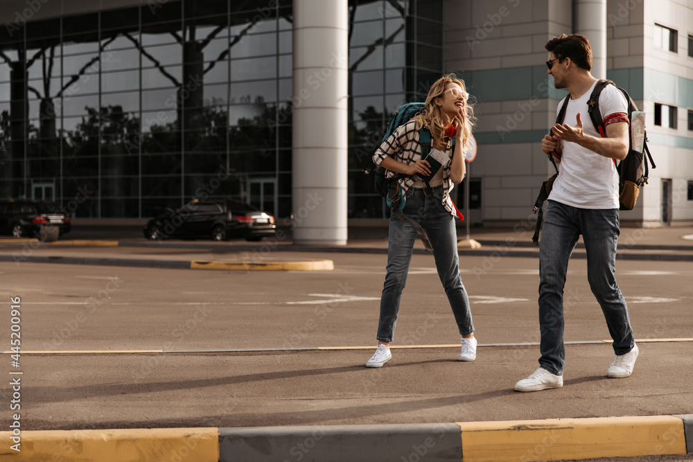 Charming woman in denim pants, plaid shirt and brunette man in jeans and white tee talks and smiles. Travelers poses with backpacks near airport.