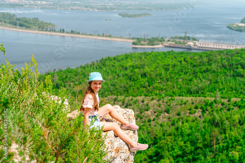 A little girl is sitting on a cliff in the mountains in summer in front of a picturesque landscape with the Volga River