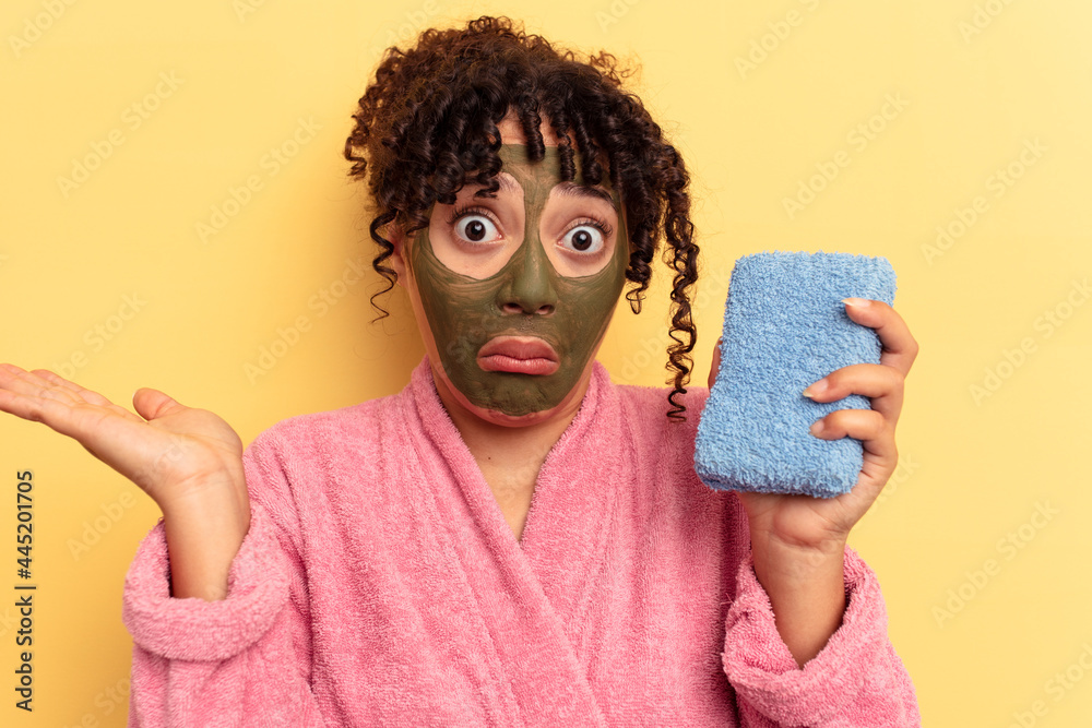 Young mixed race woman wearing a pink bathrobe holding a shower sponge isolated on yellow background shrugs shoulders and open eyes confused. Young mixed race woman wearing a pink bathrobe holding a shower sponge isolated on yellow background shrugs shoulders and open eyes confused.
