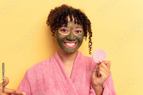 Young mixed race woman wearing a bathrobe holding a make-up remover sponge isolated on yellow background person pointing by hand to a shirt copy space, proud and confident