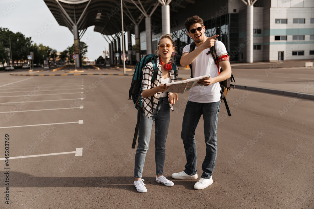 Mixed-up man and woman with map decide where to go. Guy in white t-shirt and girl in sunglasses, plaid shirt poses with backpacks near airport.