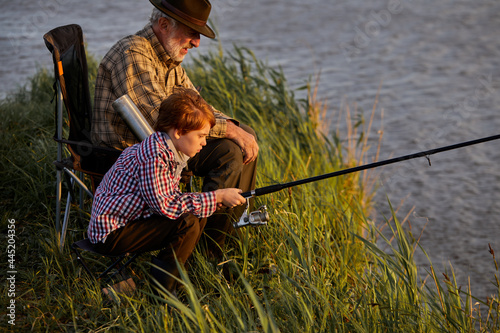 family, generation, summer holidays and people concept.happy caucasian grandfather and grandson with fishing rods on river berth. side view, copy space. weekends in nature