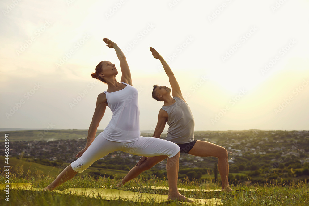 Middle aged male instructor teaching female student to do Exalted Crescent Lunge while practicing yoga on countryside meadow on peaceful summer morning. Couple doing Exalted Warrior asana in nature Middle aged male instructor teaching female student to do Exalted Crescent Lunge while practicing yoga on countryside meadow on peaceful summer morning. Couple doing Exalted Warrior asana in nature