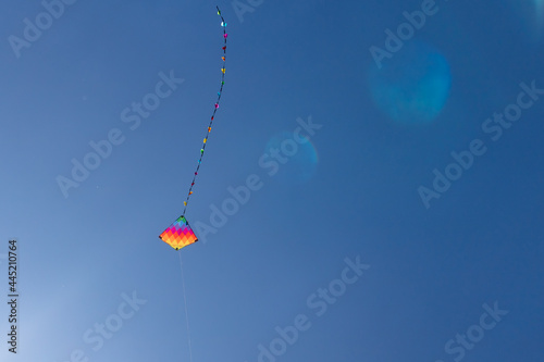 Child's rainbow coloured diamond kite with tail flying high in clear blue summer sky