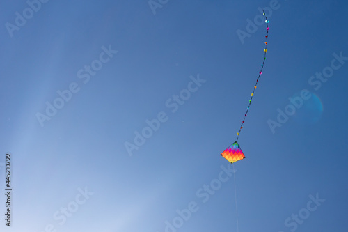 Child's rainbow coloured diamond kite with tail flying high in clear blue summer sky