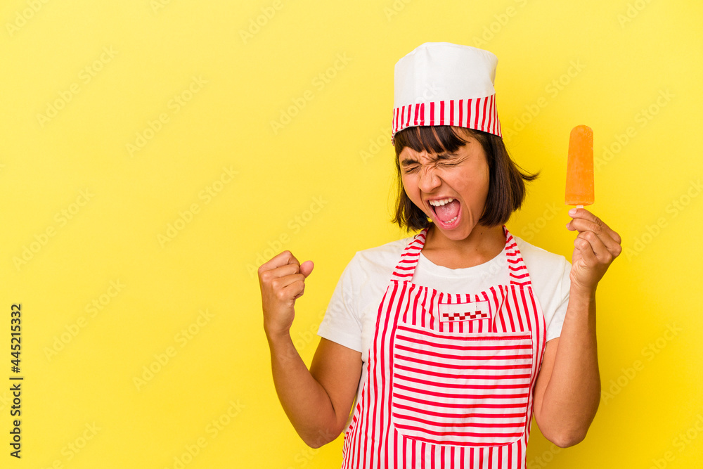 Young mixed race ice cream maker woman holding an ice cream isolated on yellow background raising fist after a victory, winner concept. Young mixed race ice cream maker woman holding an ice cream isolated on yellow background raising fist after a victory, winner concept.