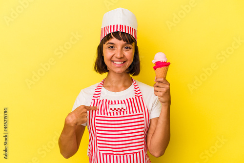 Young mixed race ice cream maker woman holding an ice cream isolated on yellow background person pointing by hand to a shirt copy space, proud and confident © Asier