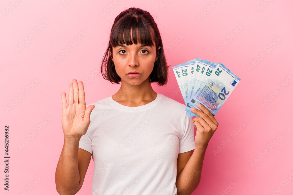 Young mixed race woman holding bills isolated on pink background standing with outstretched hand showing stop sign, preventing you. Young mixed race woman holding bills isolated on pink background standing with outstretched hand showing stop sign, preventing you.