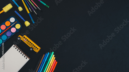 School bus and art supplies on black. School bus, paints, pencils, brushes and markers lie on the left against a black background with space for text on the right, top view close-up.