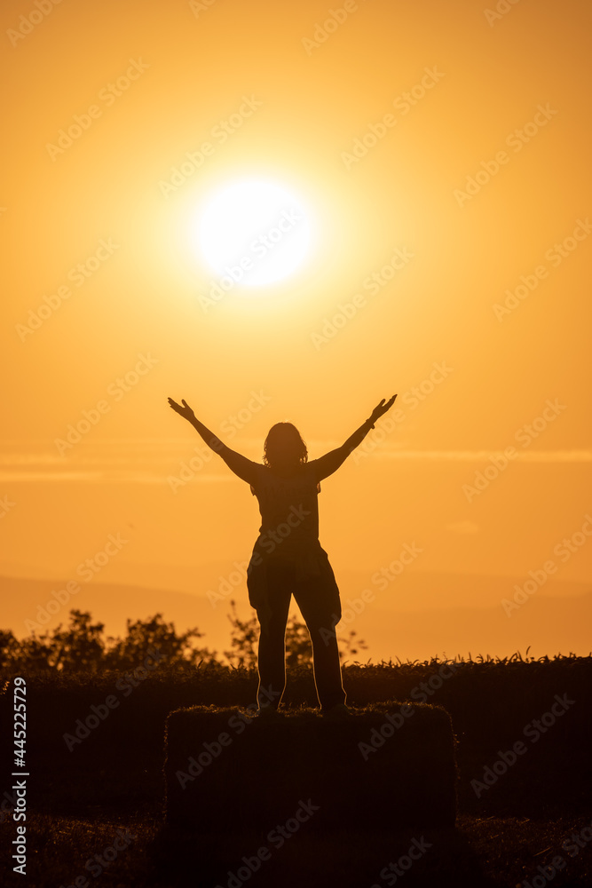 backlit photograph of the silhouette of a woman standing on a straw bale with her arms outstretched and the sun behind her