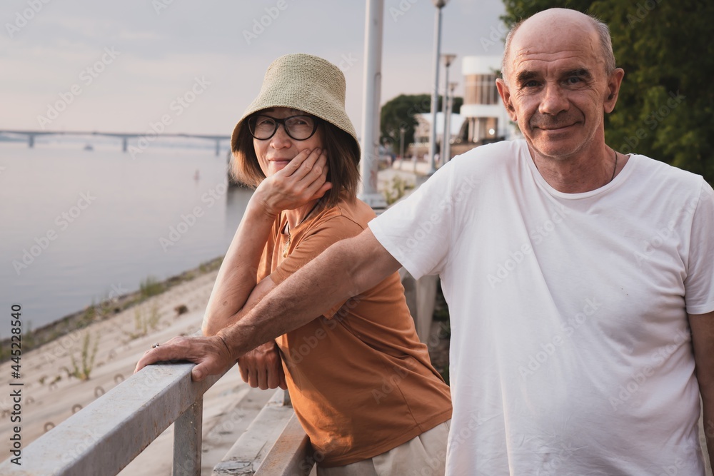 happy smiling senior couple standing on pier of river in summer vacation. people look at camera