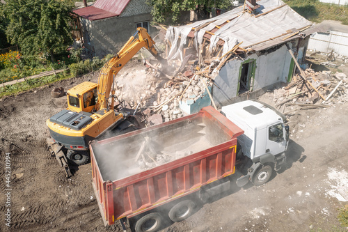 Excavator loads construction waste into truck for removal from construction site. Demolition of dilapidated housing for new development, aerial view.