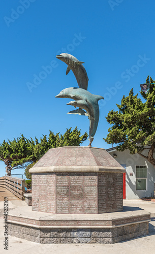 Cayucos, CA, USA - June 10, 2021: Jumping dolphins statue on brown stone pedestal with circular bench at base of pier against blue sky. Green foliage and white bird poop on dolphins. © Klodien