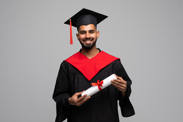 education, graduation and people concept - happy smiling indian male graduate student in mortar board and bachelor gown with diploma over grey background