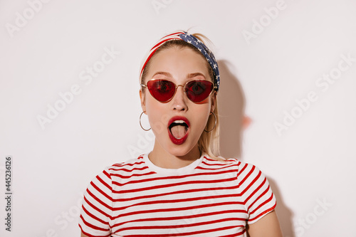 Shocked woman with red lips in sunglasses, cool earrings and striped modern t-shirt looking into camera on isolated backdrop..