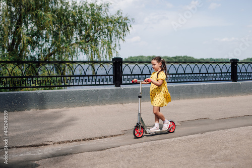 a girl in a yellow dress learns to ride a scooter given for her birthday along the city lake. 
