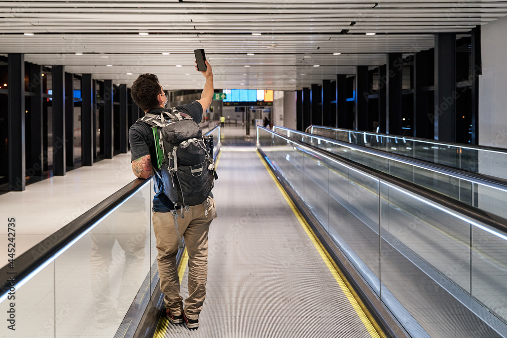 Obraz premium latin male tourist in mask advancing on airport treadmill with social distancing while making a video call during coronavirus pandemic or covid19 virus, concept of new normal