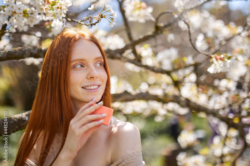 Beautiful sexy sensual woman with fresh health skin holding cream bottle in hands  in nature at spring  near blooming trees  sunny day. skincare  spa  natural cosmetic product concept