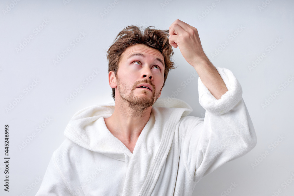 shaggy young caucasian guy in white bathrobe isolated on white background, touches long regrown hair and looks up with sad face, thinking. Beauty, people lifestyle concept. Copy space. Hairstyle shaggy young caucasian guy in white bathrobe isolated on white background, touches long regrown hair and looks up with sad face, thinking. Beauty, people lifestyle concept. Copy space. Hairstyle