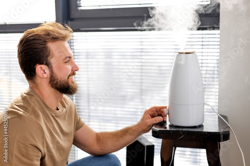 Young smiling bearded male turn on purifier in room, in smart house, side view portrait. Air cleaning and humidifying device for house. Modern technologies, innovation concept. Focus on man photo