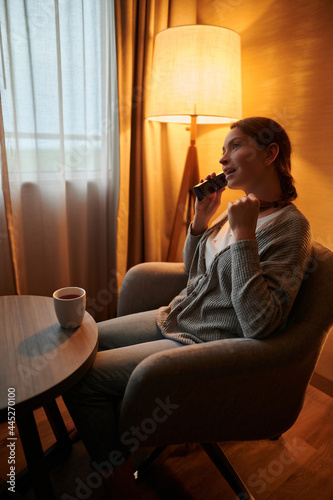 young attractive woman is sitting on the sofa in the room, emotionally talking on the phone. There is a floor lamp in the corner of the room. There is a cup of tea on the table.