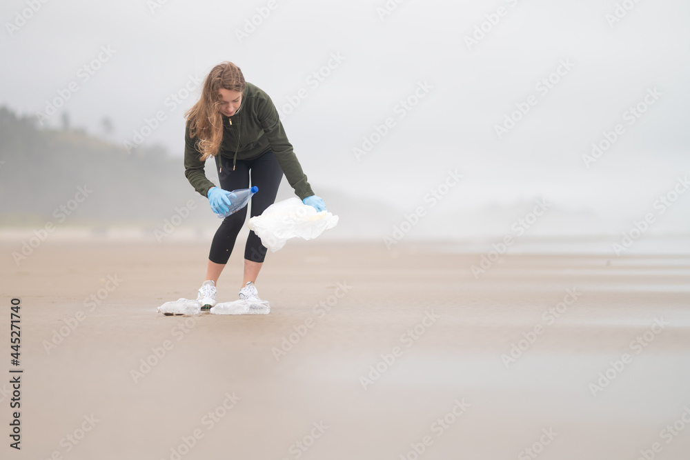 A woman volunteer collects garbage in a bag on the ocean shore. Cool day. The woman is wearing a jacket. She has long blonde hair. Minimalism. Empty space for your insert.