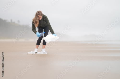 A woman volunteer collects garbage in a bag on the ocean shore. Cool day. The woman is wearing a jacket. She has long blonde hair. Minimalism. Empty space for your insert.