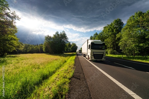 Obraz na płótnie Two white trucks driving on the asphalt road between the forest and the glade at sunset w salonie