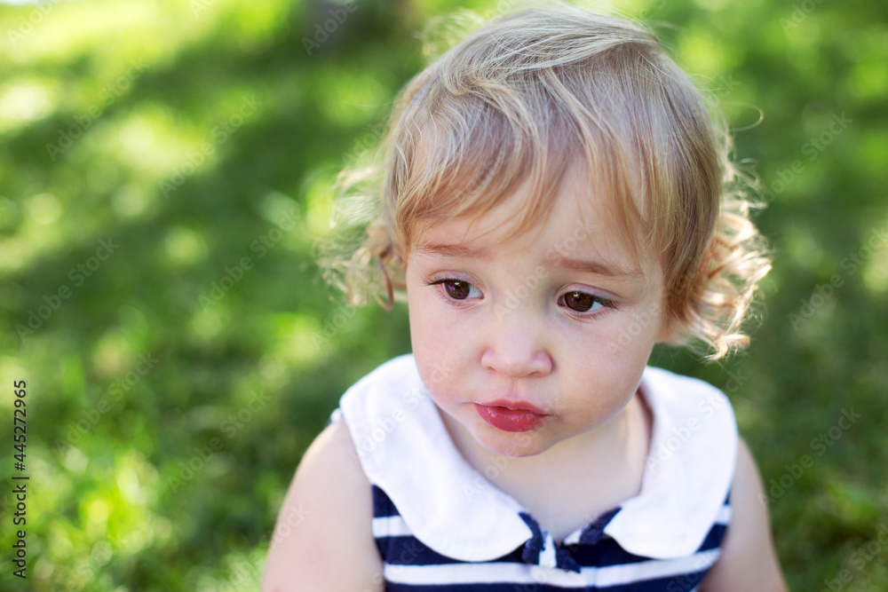 Portrait of cute curly carefree surprised toddler girl outdoors in sunny summer day against green grass
