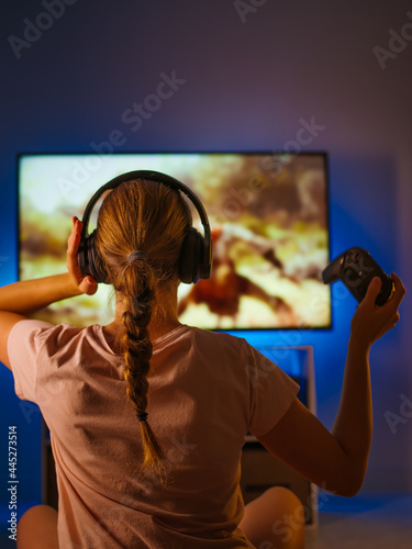 A young woman remotely plays a video game. She holds a joystick in her hand, and has headphones on her head. In front of the woman is a large computer monitor. Youth culture, video games.
