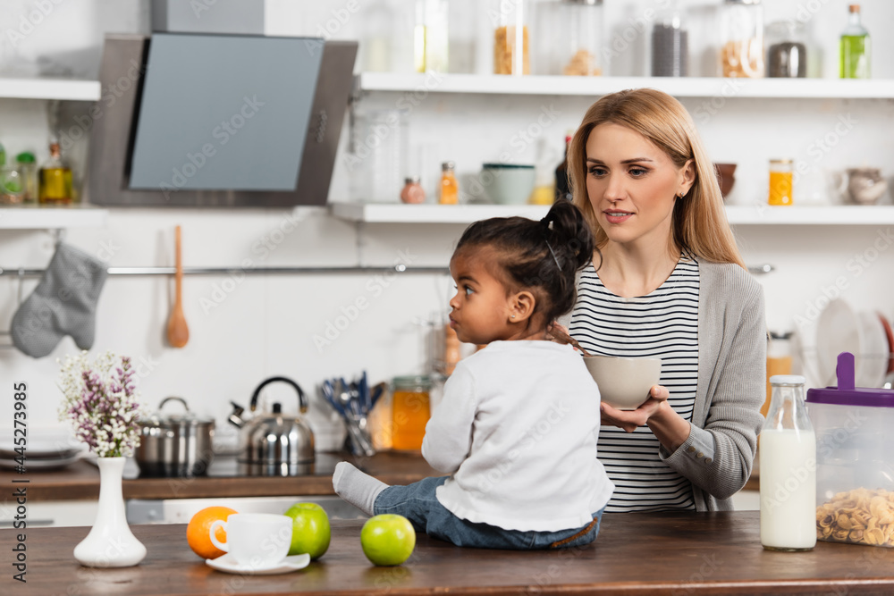 woman holding bowl and looking at adopted african american kid sitting on kitchen table